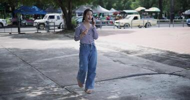 Handheld shot, A cheerful Asian woman takes a leisurely stroll along a concrete walkway while talking with friend on smartphone in a park. Weekend relaxation time video