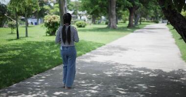 Handheld back view shot, A cheerful Asian woman enjoys take a photo by camera and takes a leisurely stroll along a concrete walkway in a park. Weekend relaxation time video