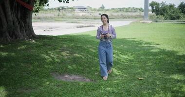 Handheld shot, Cheerful woman is walking in a park on a sunny day, surrounded by lush green trees, and enjoys taking pictures of the nature with her camera. A relaxing weekend moment. video