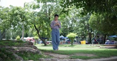 Handheld shot, Happy young woman playing with her mobile phone while taking a leisurely stroll, surrounded by lush green trees in a park. Enjoy holiday activities video