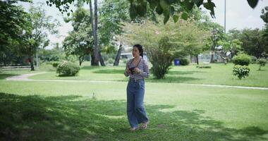 Handheld shot, Cheerful woman is walking in a park on a sunny day, surrounded by lush green trees, and enjoys taking pictures of the nature with her camera. A relaxing weekend moment. video