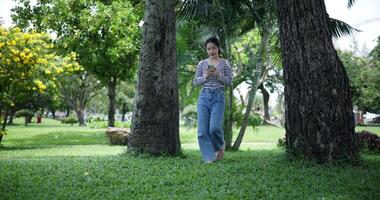 Handheld shot, Happy young woman playing with her mobile phone while taking a leisurely stroll, surrounded by lush green trees in a park. Enjoy holiday activities video