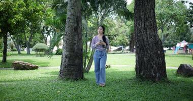 Handheld shot, Happy young woman playing with her mobile phone while taking a leisurely stroll, surrounded by lush green trees in a park. Enjoy holiday activities video