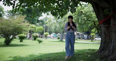 Handheld shot, Cheerful woman is walking in a park on a sunny day, surrounded by lush green trees, and enjoys taking pictures of the nature with her camera. A relaxing weekend moment. video