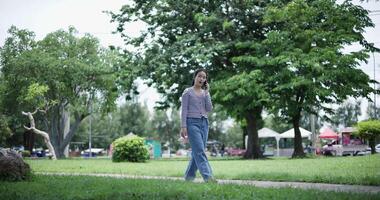 Handheld shot, A cheerful Asian woman takes a leisurely stroll along a concrete walkway while talking with friend on smartphone in a park. Weekend relaxation time video