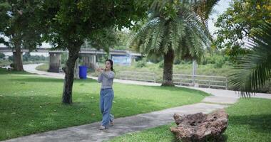 Handheld shot, Happy young woman is talking with her friend via call on smartphone while taking a leisurely stroll, surrounded by lush green trees in a park. Enjoy holiday activities video