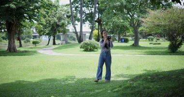 Handheld shot, Cheerful woman is walking in a park on a sunny day, surrounded by lush green trees, and enjoys taking pictures of the nature with her camera. A relaxing weekend moment. video