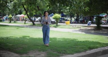 Handheld shot, Cheerful woman is walking in a park on a sunny day, surrounded by lush green trees, and enjoys taking pictures of the nature with her camera. A relaxing weekend moment. video