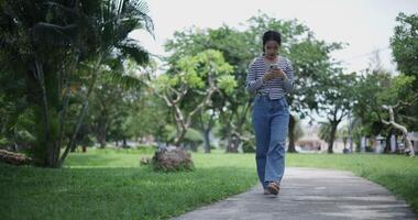 Handheld shot, Happy young woman playing with mobile phone while leisurely walking on concrete walkway in park. Weekend relaxation time video