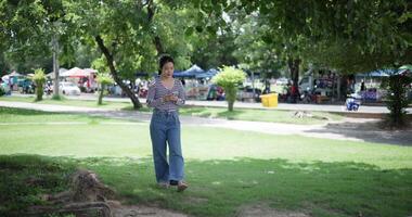 Handheld shot, Happy young woman playing with her mobile phone while taking a leisurely stroll, surrounded by lush green trees in a park. Enjoy holiday activities video