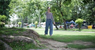 Handheld shot, Happy young woman is talking with her friend via call on smartphone while taking a leisurely stroll, surrounded by lush green trees in a park. Enjoy holiday activities video