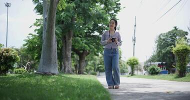 Handheld shot, A cheerful Asian woman enjoys take a photo by camera and takes a leisurely stroll along a concrete walkway in a park. Weekend relaxation time video