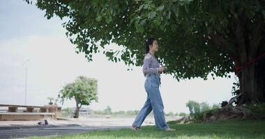 Handheld side view shot, Cheerful woman is walking in a park on a sunny day, surrounded by lush green trees, and enjoys taking pictures of the nature with her camera. A relaxing weekend moment. video