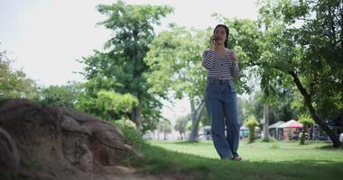 Handheld shot, Happy young woman is talking with her friend on smartphone while taking a leisurely stroll, surrounded by lush green trees in a park. Enjoy holiday activities video