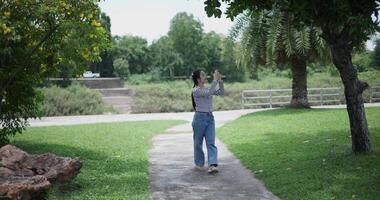 Handheld shot, Cheerful woman is walking in a park on a sunny day, surrounded by lush green trees, and enjoys taking pictures of the nature with smartphone. A relaxing weekend moment. video