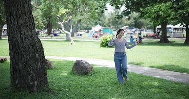 Handheld shot, Cheerful woman wearing headphones is walking in a park, happily dancing and raises her hands up, feeling relaxed and carefree. video