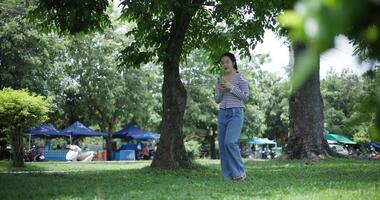 Handheld shot, Happy young woman playing with her mobile phone while taking a leisurely stroll, surrounded by lush green trees in a park. Enjoy holiday activities video