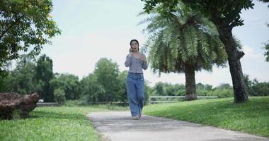 Handheld shot, A cheerful Asian woman takes a leisurely stroll along a concrete walkway while talking with friend on smartphone in a park. Weekend relaxation time video