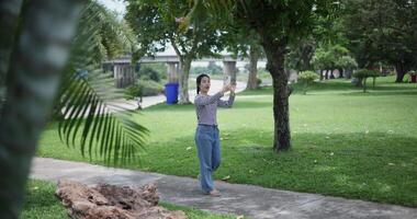 Handheld shot, Happy young woman is talking with her friend via call on smartphone while taking a leisurely stroll, surrounded by lush green trees in a park. Enjoy holiday activities video