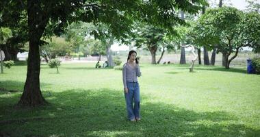 Handheld shot, Happy young woman is talking with her friend on smartphone while taking a leisurely stroll, surrounded by lush green trees in a park. Enjoy holiday activities video