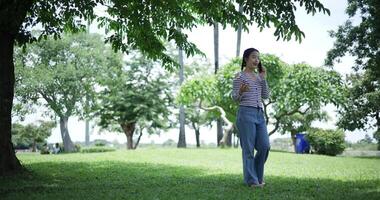 Handheld shot, Happy young woman is talking with her friend on smartphone while taking a leisurely stroll, surrounded by lush green trees in a park. Enjoy holiday activities video