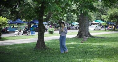Handheld shot, Happy young woman playing with her mobile phone while taking a leisurely stroll, surrounded by lush green trees in a park. Enjoy holiday activities video
