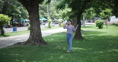Handheld shot, Happy young woman is talking with her friend via call on smartphone while taking a leisurely stroll, surrounded by lush green trees in a park. Enjoy holiday activities video
