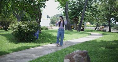Handheld shot, Cheerful asian woman using mobile phone while enjoys music from wireless headphones, singing and dancing in a park, feeling relaxed and carefree. video