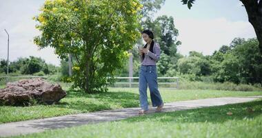 Handheld shot, Cheerful asian woman leisurely strolling and using smartphone while enjoying music from wireless headphones with fresh air in a park. feeling relaxed and carefree. video