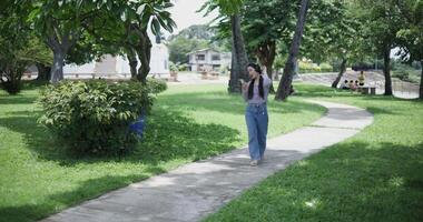 Handheld shot, Cheerful asian woman using mobile phone while enjoys music from wireless headphones, singing and dancing in a park, feeling relaxed and carefree. video