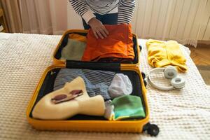 Open yellow suitcase with different family clothes and accessories on sofa. Woman packing suitcase for trip on floor indoors, close up photo