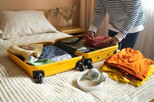 Open yellow suitcase with different family clothes and accessories on sofa. Woman packing suitcase for trip on floor indoors, close up photo