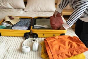 Open yellow suitcase with different family clothes and accessories on sofa. Woman packing suitcase for trip on floor indoors, close up photo