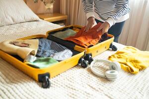 Open yellow suitcase with different family clothes and accessories on sofa. Woman packing suitcase for trip on floor indoors, close up photo