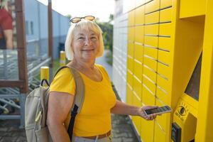 Happy smiling 50s senior woman client with smart phone at outdoor automated parcel machine hoosing operation he touch screened on smart background photo
