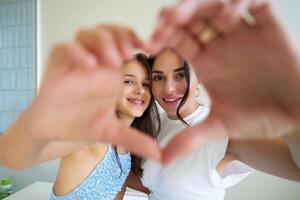 A mother and daughter making a heart shape with their hands while smiling at each other indoors photo