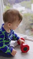 Little adorable baby boy playing with a red dumb-bell on the floor. Lovely toddler crawling by the floor at big panoramic window. High angle view. Vertical video