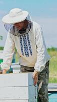a beekeeper in protective clothing holds a frame with honeycombs for bees in the garden in the summer Vertical video