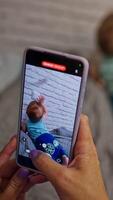 Mommy is taking of her son on the smartphone. Little baby boy crawling by the bed exploring the white brick wall. Blurred backdrop. Vertical video