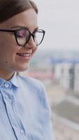 Happy positive smiling woman standing at the big panoramic window. Businesswoman taking notes into paper book and then looking into window. Cityscape at backdrop in blur. Vertical video
