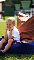 Smiling beautiful kid sits in a comfortable bean bag chair outdoors. Approaching blond boy spending time in resort zone. Vertical video
