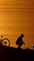 Man with bicycle resting on beach. Silhouette of man resting after cycling trip near river Vertical video