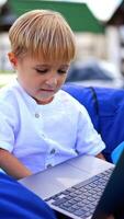 Happy little Caucasian boy watching something on computer. Blond kid smiling happily sitting in a chair in nature. Blurred backdrop. Vertical video