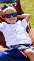 Smiling blond little boy enjoying his time in bean bag chair outdoors. Child in summer clothes and sunglasses relaxing and sunbathing. Vertical video