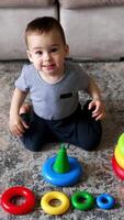 Active Caucasian toddler jumps on the floor. Toy pyramids on the floor in front of baby. High angle view. Vertical video