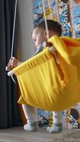 Adorable baby boy swinging in the swing at home and smiles happily. Black cat sits on the floor beside. Low angle view. Vertical video