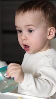 Little toddler sitting at the feeding table reaches hand for the bottle. Mom's hand gives a bottle, kid drinks water and puts it on the table. Close up. Vertical video