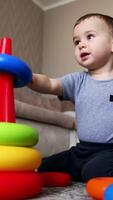 Joyful baby boy playing with toy pyramid. Kid takes two toy rings and bangs them together. Low angle view. Vertical video