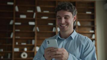 Happy wide smiling young Caucasian male business man businessman guy entrepreneur using mobile phone smartphone typing message chatting browsing internet enjoy online app game ordering delivery indoor video
