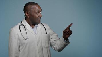 African American man doctor medical worker in white coat male healthcare employee medic customer in studio blue background pointing index finger making choice choosing smiling good selection medicare video